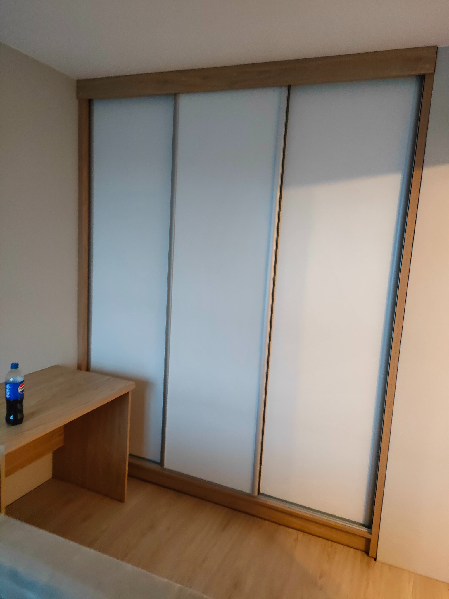 Jasna szafa wnękowa z przesuwnymi drzwiami i drewnianą ramą, z blatem po lewej stronie, na którym stoi butelka napoju. Minimalistyczny design.