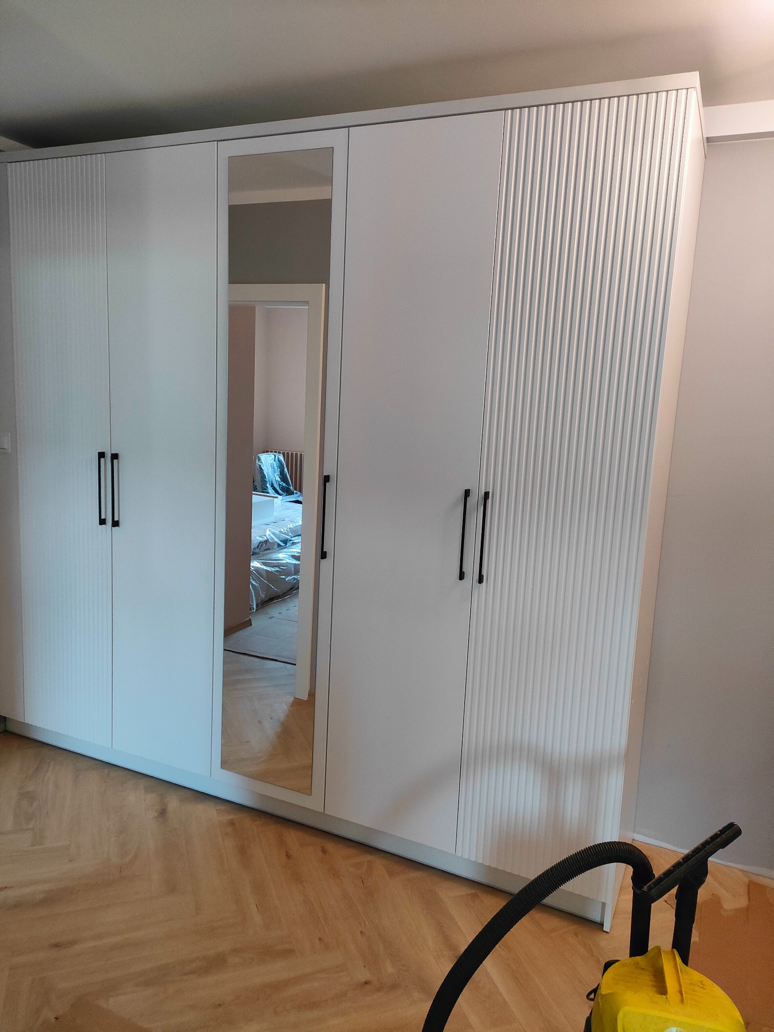 Biała szafa wnękowa z lustrem i frezowanymi frontami, czarne podłużne uchwyty, parkiet w jodełkę, widoczny odkurzacz. Wnętrze pokoju widoczne w lustrze.