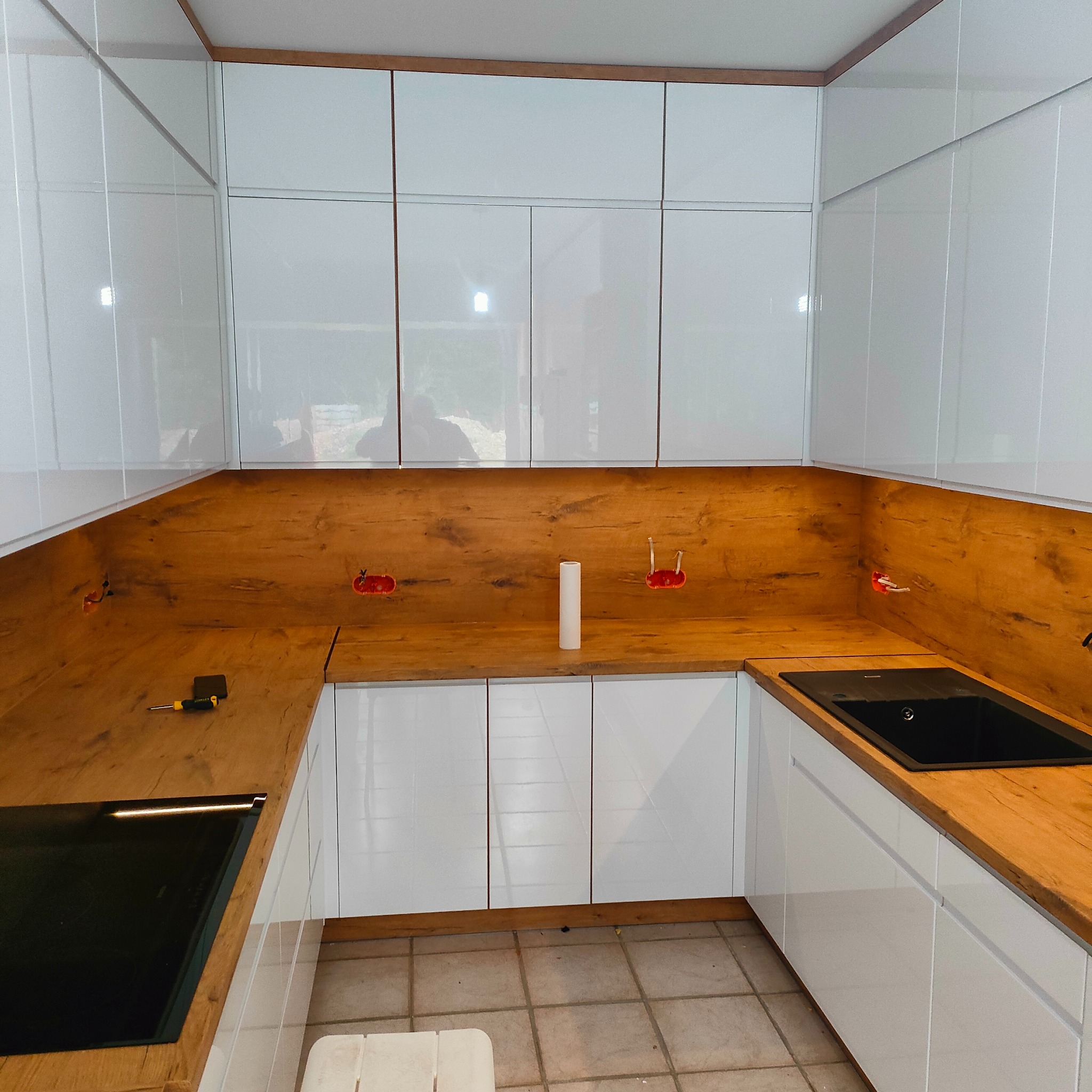 Kątowe ujęcie kuchni z białymi, lakierowanymi szafkami i drewnianym blatem. Widoczne gniazdka elektryczne, zlewozmywak i płyta indukcyjna. Na blacie leży śrubokręt.