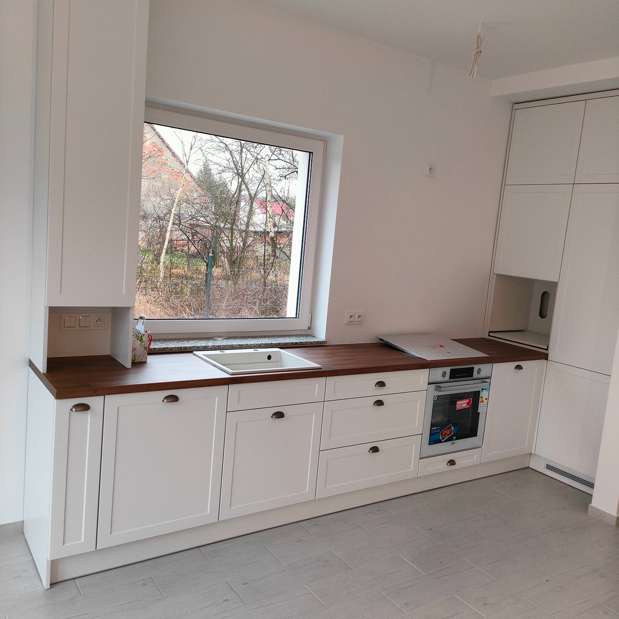 Jasna kuchnia z drewnianym blatem i wbudowanym oknem z widokiem na drzewa. Uwagę przykuwają metalowe uchwyty i minimalistyczny design.