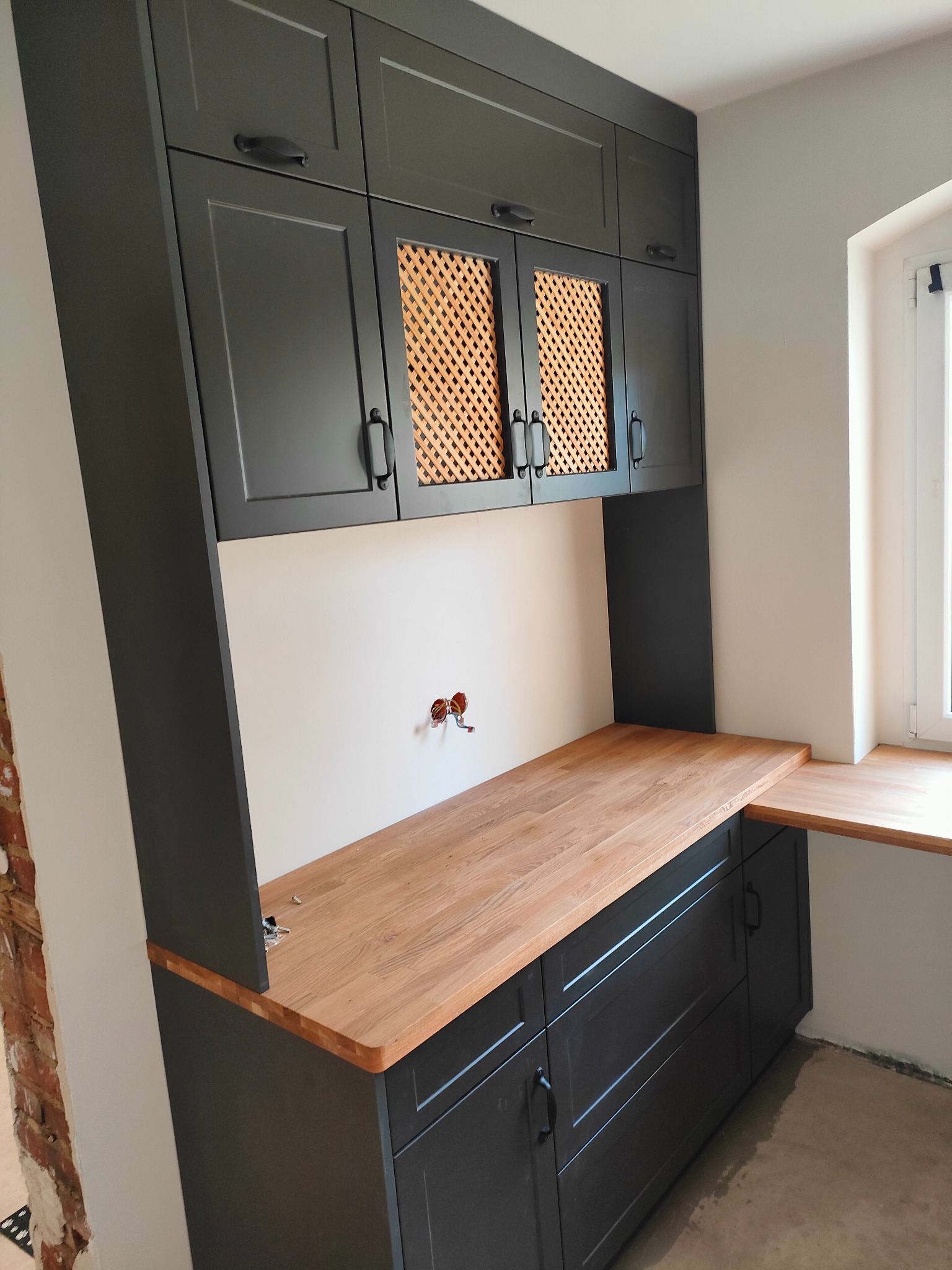 Antracytowa zabudowa kuchenna z drewnianym blatem i ażurowymi frontami szafek wiszących. Widoczne gniazdka elektryczne i fragment ściany z cegły.