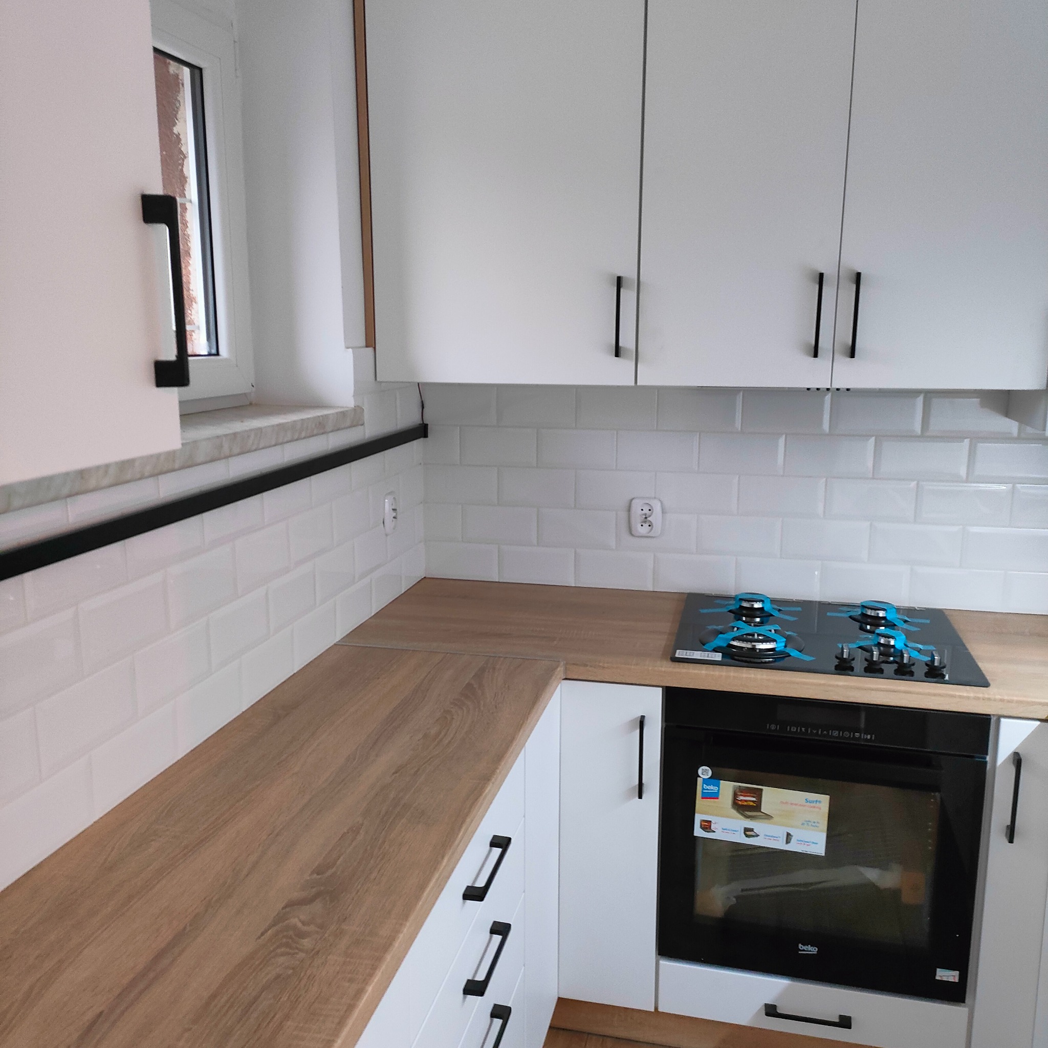 Biała kuchnia z drewnianym blatem, czarnymi uchwytami i płytkami na ścianie. Widoczny piekarnik i płyta gazowa. Nowoczesny design, jasne wnętrze.