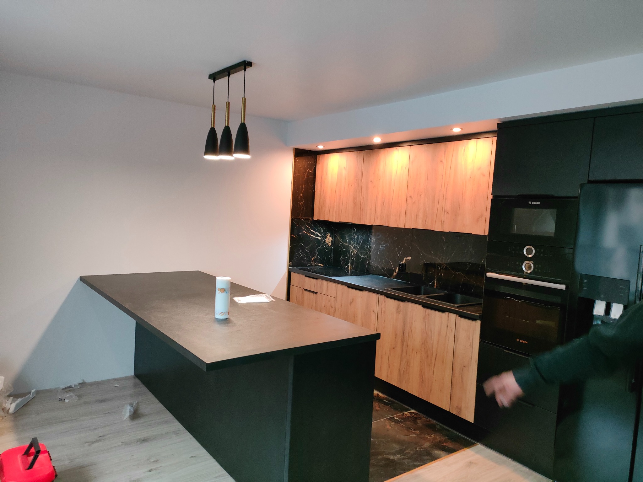 Nowoczesna kuchnia z wyspą: drewniane fronty szafek, czarny blat i marmurowy backsplash. Nad wyspą wiszą trzy czarne lampy.