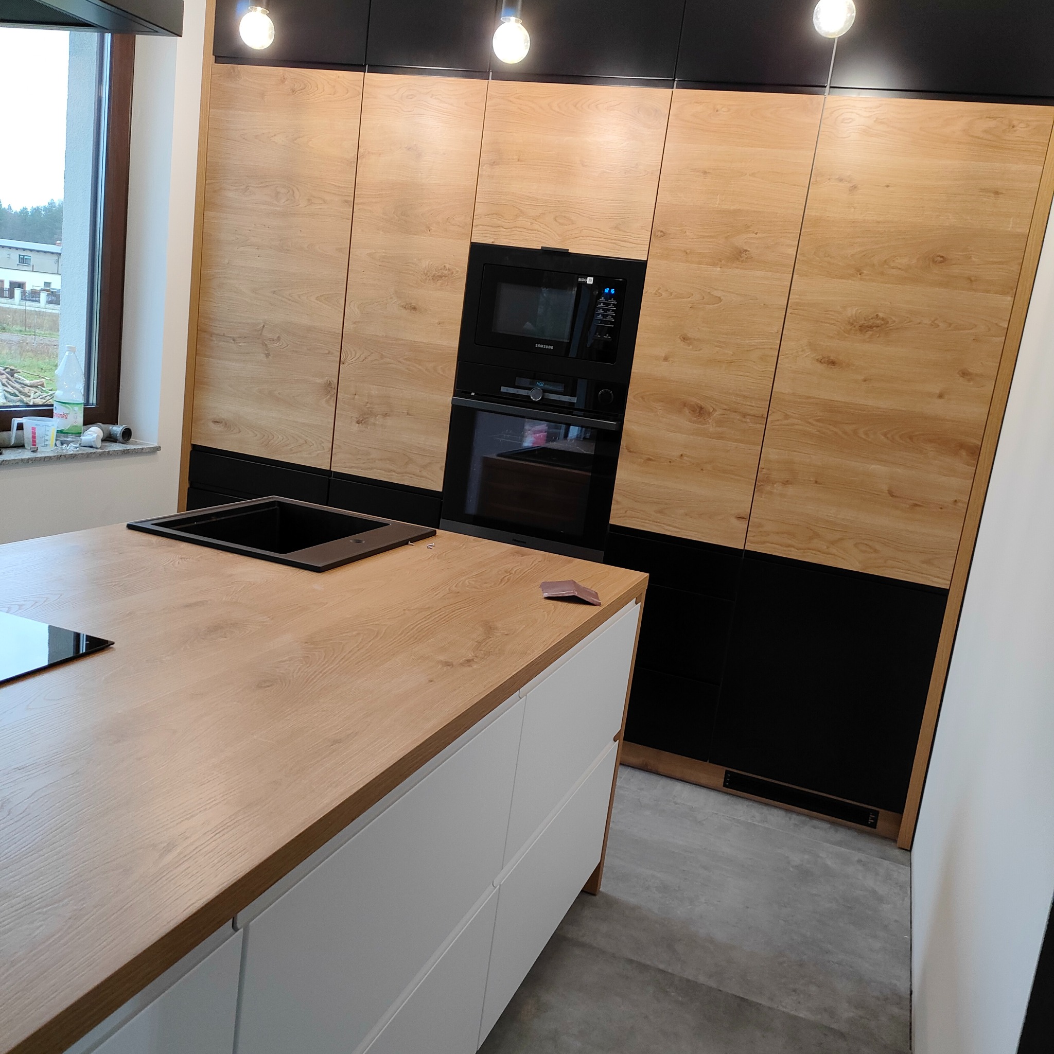 Nowoczesna kuchnia: drewniane fronty szafek, czarne akcenty, wyspa z białymi szufladami i drewnianym blatem. Widoczny zlew i płyta indukcyjna.