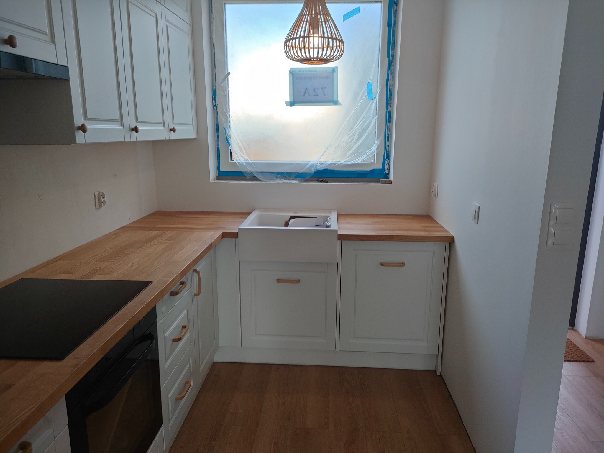 Jasna kuchnia z drewnianym blatem i białymi szafkami, okno zabezpieczone folią, lampa wisząca. Widok na fragment płyty grzewczej i piekarnika.