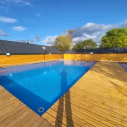 Błękitny basen otoczony drewnianym tarasem w słoneczny dzień. Widoczne odbicie osoby. Jasne deski tarasu kontrastują z ciemną elewacją budynku w tle.