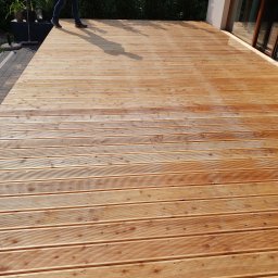 CLIC HOUSE WOOD - Nowy, drewniany taras z ryflowanej deski, z widocznymi śladami świeżego lakierowania, częściowo w cieniu osoby stojącej na jego skraju. Przy budynku widoczne przesuwne drzwi.