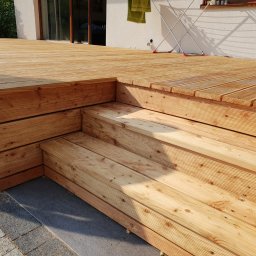 CLIC HOUSE WOOD - Jasny taras z desek z niskimi schodami, oświetlony słońcem. W tle biały budynek z suszarką na pranie. Widoczne detale łączeń desek i faktura drewna.