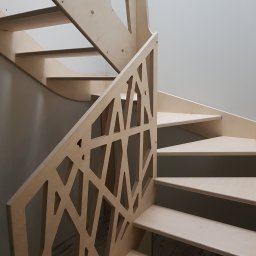 CLIC HOUSE WOOD - Spiralne, jasne drewniane schody z ażurową balustradą o geometrycznym wzorze, widoczne z dołu. Naturalne oświetlenie, minimalistyczny design.