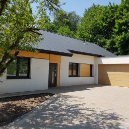 CLIC HOUSE WOOD - Nowoczesny dom jednorodzinny z szarą blachodachówką i elementami drewnianymi na elewacji, otoczony zielenią. Widoczny podjazd z kostki brukowej i drzewo z lewej strony.