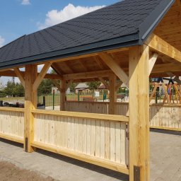 CLIC HOUSE WOOD - Drewniana altana z czarnym dachem na tle placu zabaw i błękitnego nieba. Widoczne jasne drewniane elementy konstrukcyjne i wypełnienie ścianek.