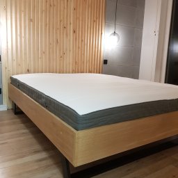 CLIC HOUSE WOOD - Drewniane łóżko z materacem na tle ściany z drewnianych paneli i betonowych płyt. Nad łóżkiem wisi szklana lampa. Minimalistyczny design sypialni.