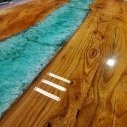 CLIC HOUSE WOOD - Błyszczący blat stołu z drewna z żywicą w kolorze turkusowym imitującą rzekę, widoczne słoje drewna i refleksy świetlne, całość w ciepłej tonacji.