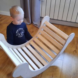 CLIC HOUSE WOOD - Jasnowłosy chłopiec bawi się drewnianą, bujaną konstrukcją na podłodze. Naturalne światło, widoczny grzejnik i zasłona w tle.