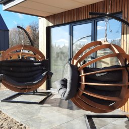 CLIC HOUSE WOOD - Dwa designerskie, drewniane fotele wiszące typu kokon z poduszkami na tarasie z płyt, na tle drewnianej elewacji budynku. Słoneczny dzień.