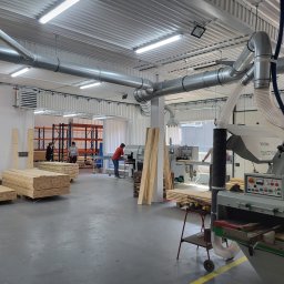 CLIC HOUSE WOOD - Wnętrze hali produkcyjnej z maszynami do obróbki drewna, ułożonymi deskami i pracownikami w tle. Widoczne rury wentylacyjne pod sufitem.