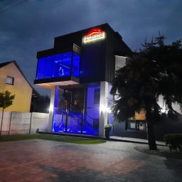 CLIC HOUSE WOOD - Nowoczesny dom z przeszkloną fasadą i podświetleniem LED w kolorze niebieskim, widoczny z zewnątrz nocą, z logo firmy na dachu.