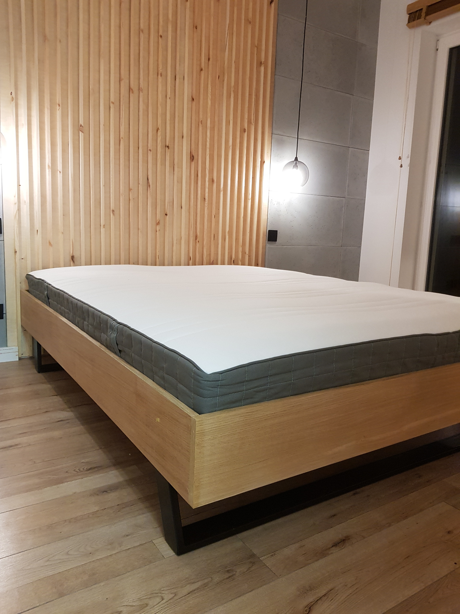 Drewniane łóżko z materacem na tle ściany z drewnianych paneli i betonowych płyt. Nad łóżkiem wisi szklana lampa. Minimalistyczny design sypialni.