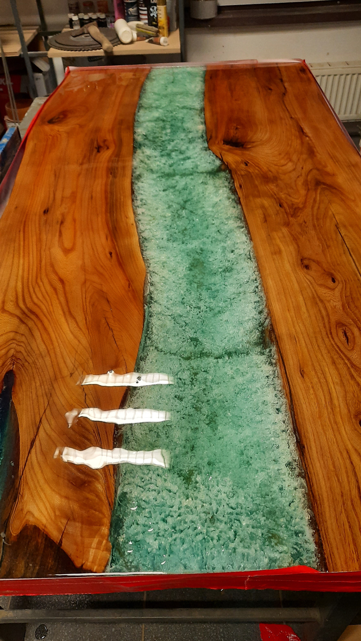 Blat stołu z drewna z żywicą epoksydową w kolorze turkusowym, imitującą rzekę. Widoczne słoje drewna i refleksy świetlne na powierzchni żywicy.