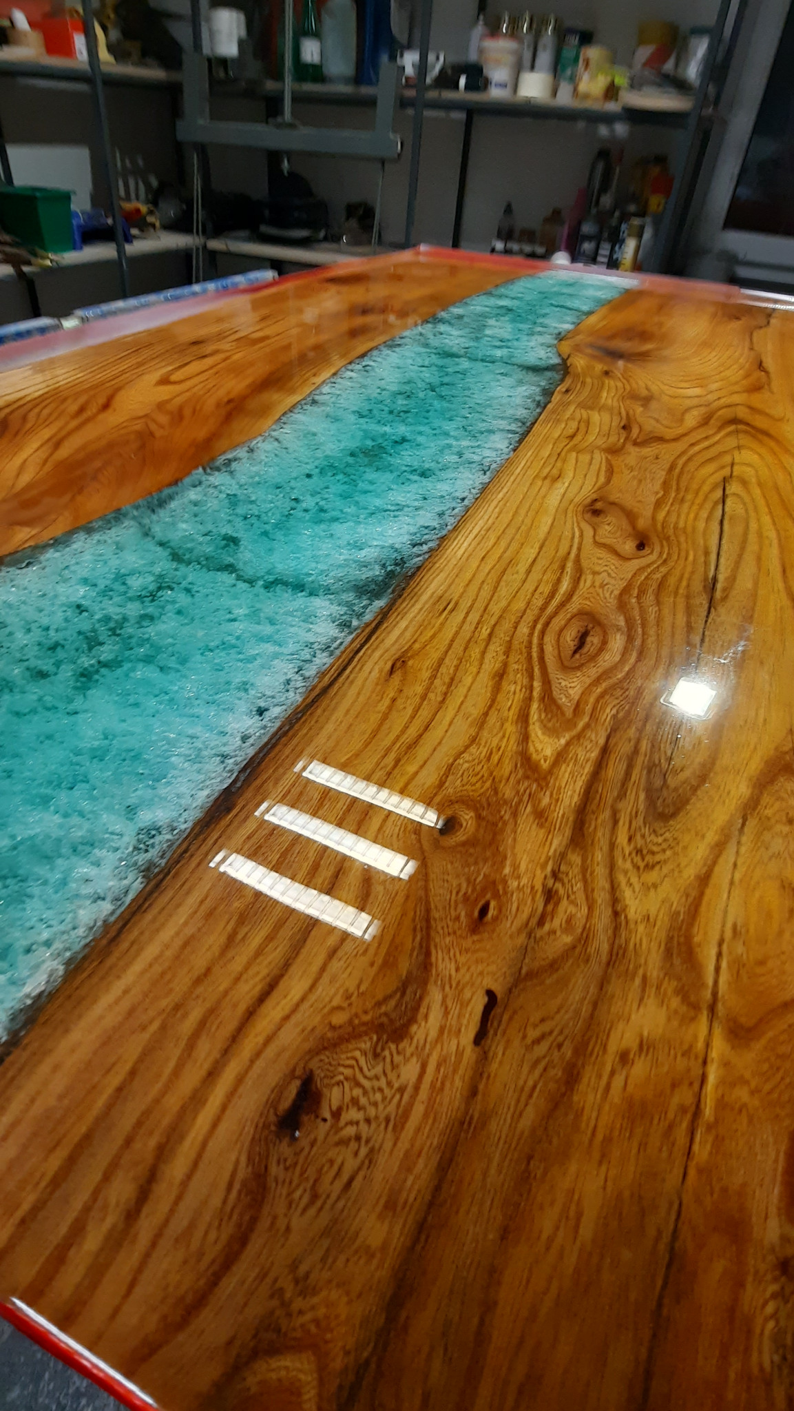 Błyszczący blat stołu z drewna z żywicą w kolorze turkusowym imitującą rzekę, widoczne słoje drewna i refleksy świetlne, całość w ciepłej tonacji.