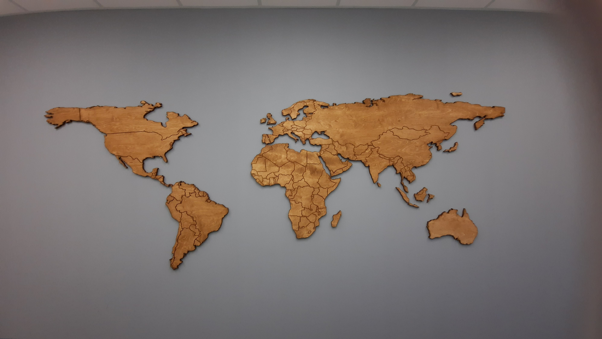 Drewniana mapa świata w odcieniach brązu, zamontowana na gładkiej, szarej ścianie. Kontynenty wycięte z drewna, z widocznymi granicami państw. Dekoracja wnętrza.