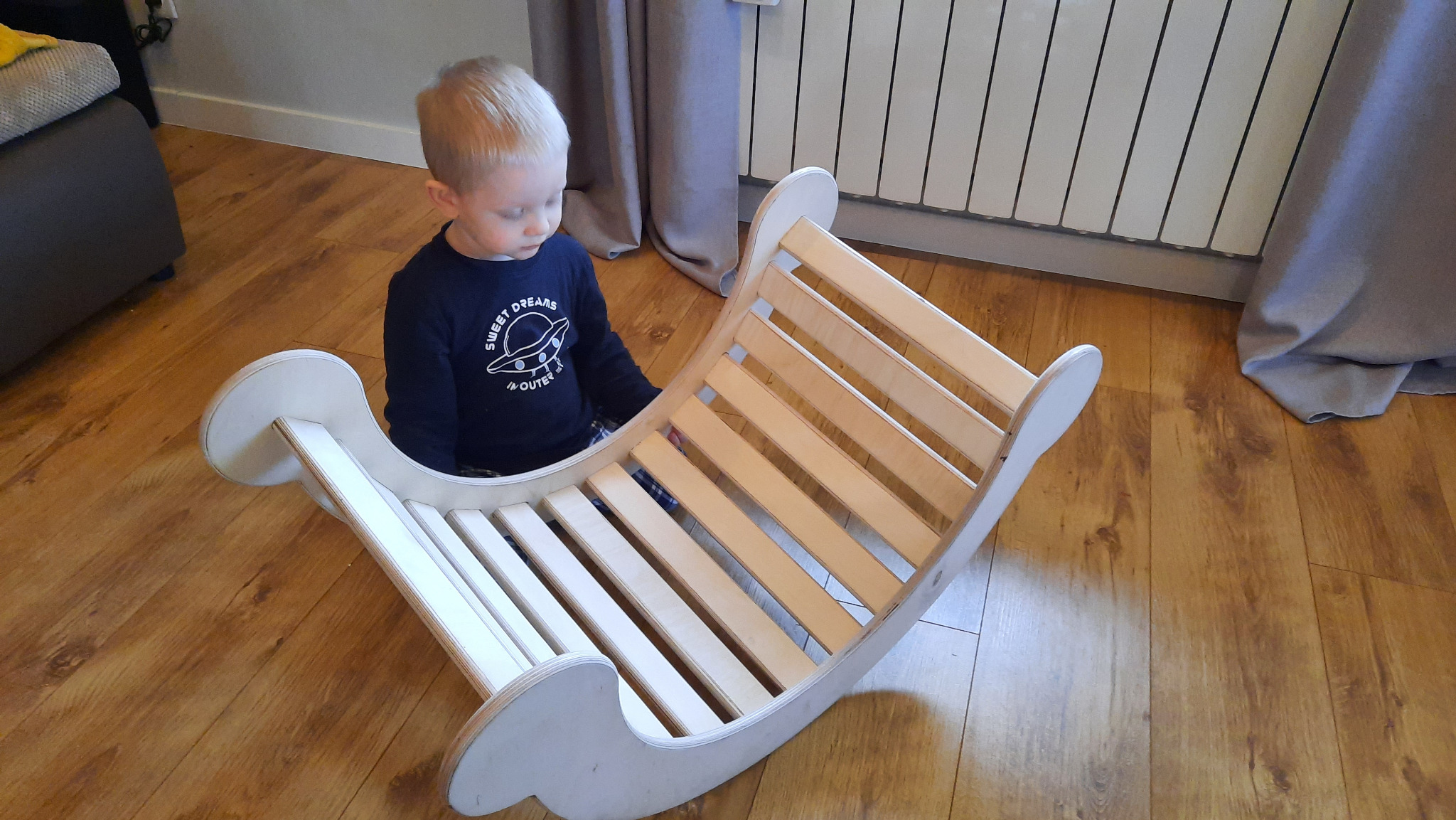 Jasnowłosy chłopiec bawi się drewnianą, bujaną konstrukcją na podłodze. Naturalne światło, widoczny grzejnik i zasłona w tle.