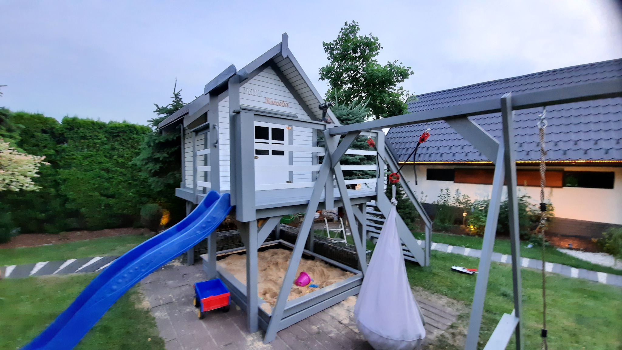 Drewniany plac zabaw dla dzieci z domkiem, zjeżdżalnią, huśtawką i piaskownicą, pomalowany na szaro i biało, otoczony zielenią. Na domku widnieje napis 'Felix i Kamelia'.