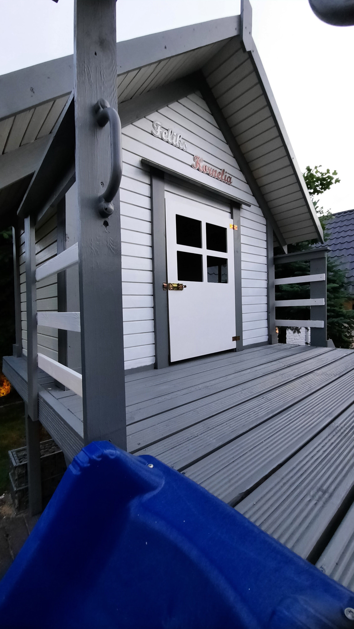 Drewniany domek dla dzieci na podeście z balustradą i zjeżdżalnią. Szara konstrukcja z białymi akcentami, imiona 'Feliks i Kornelia' nad drzwiami.