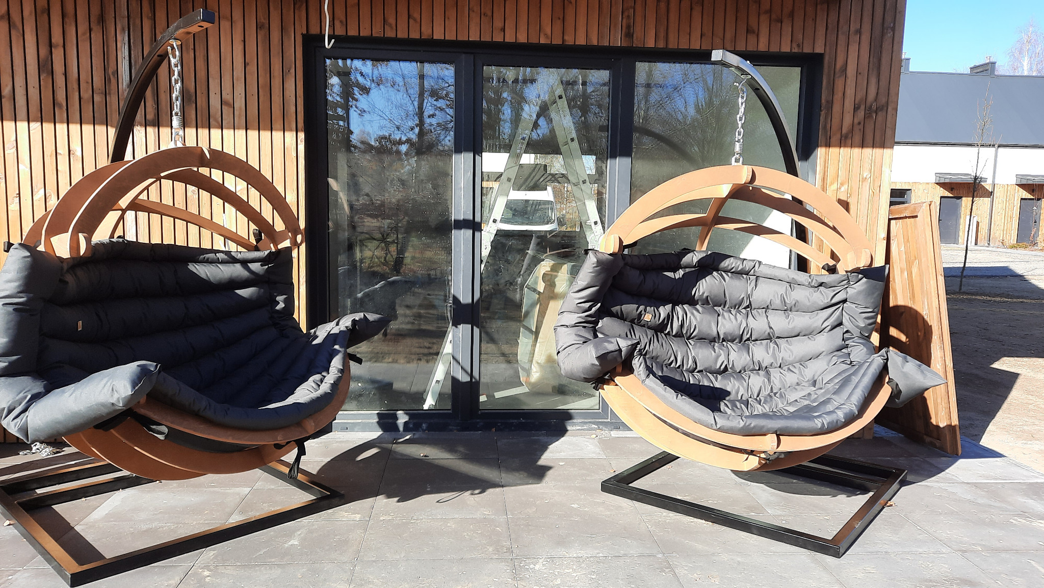 Dwa wiszące fotele z drewnianą konstrukcją i szarą poduchą na tarasie z płyt. W tle budynek z drewnianą elewacją i drabina oparta o okno.