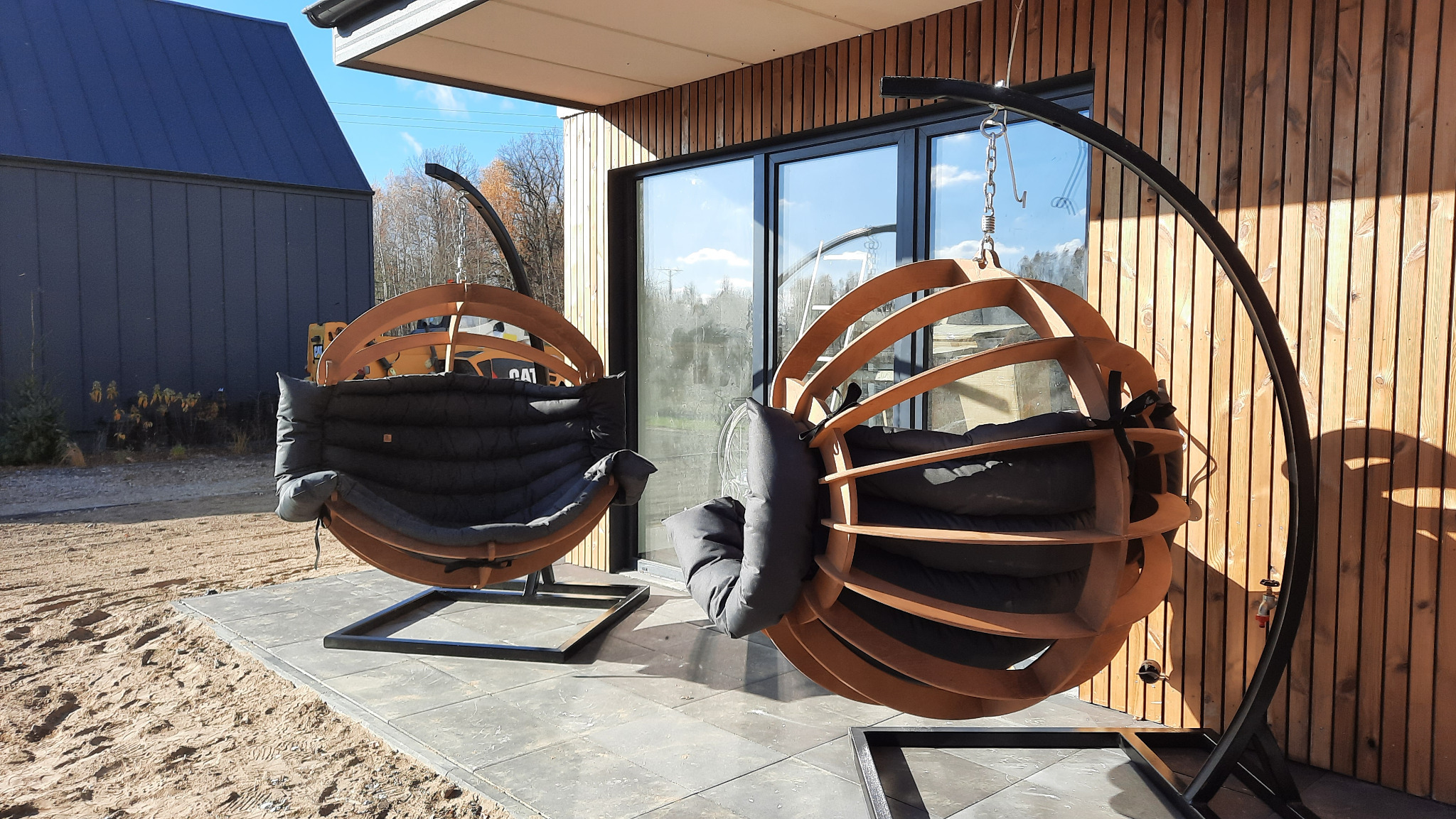 Dwa designerskie, drewniane fotele wiszące typu kokon z poduszkami na tarasie z płyt, na tle drewnianej elewacji budynku. Słoneczny dzień.