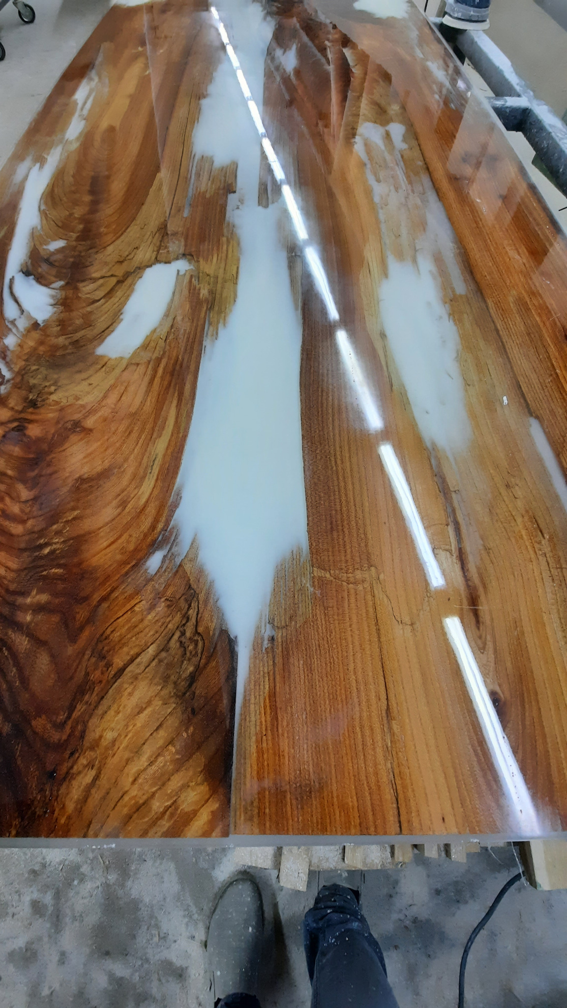 Bursztynowy blat drewniany zalany białą żywicą epoksydową, widoczne słoje drewna i refleksy światła. Nogi osoby w roboczym obuwiu w dolnej części kadru.