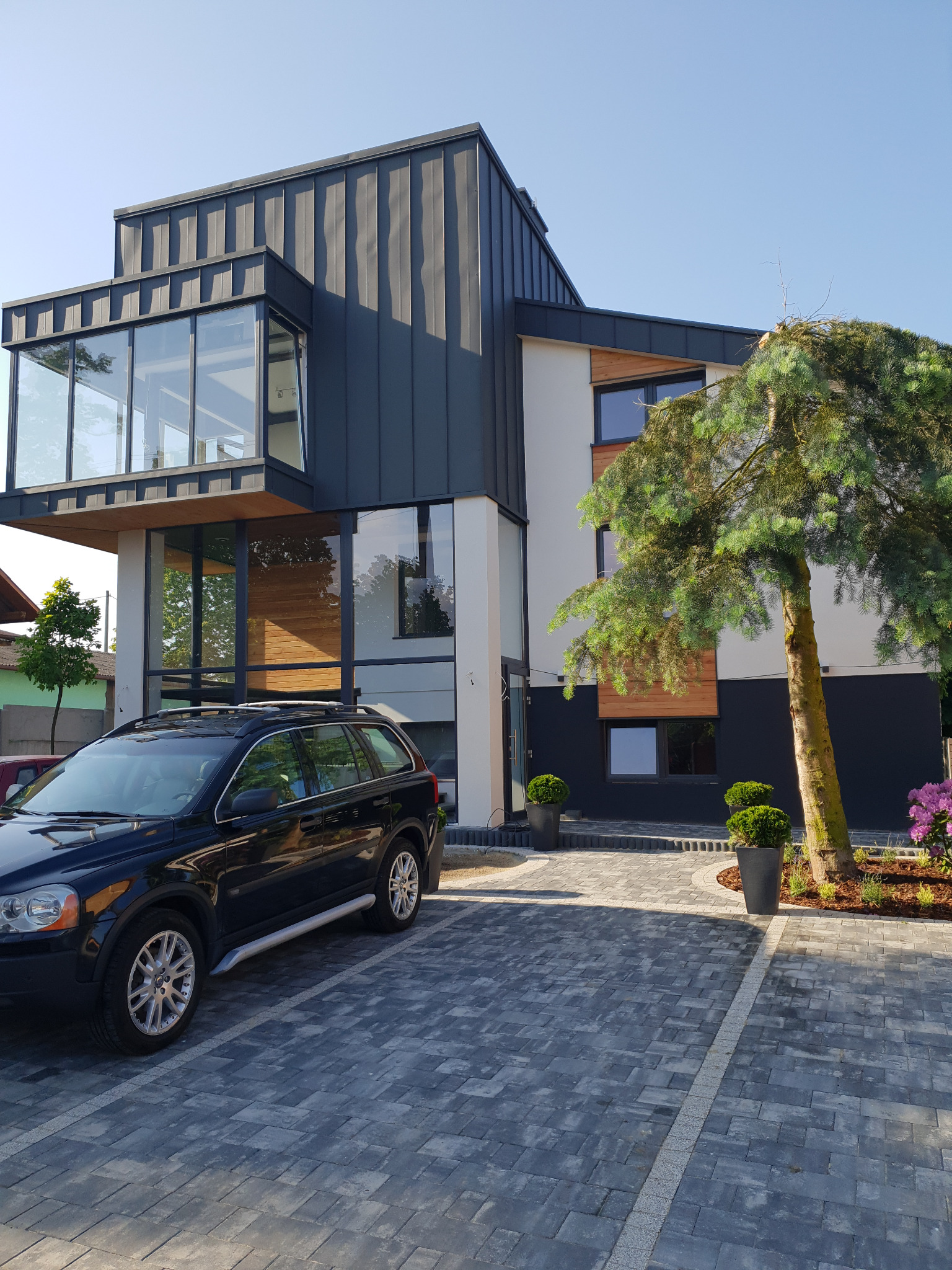 Nowoczesny dom z przeszklonym wykuszem, czarną blachą na elewacji i drewnianymi akcentami. Na pierwszym planie czarny SUV i szara kostka brukowa.