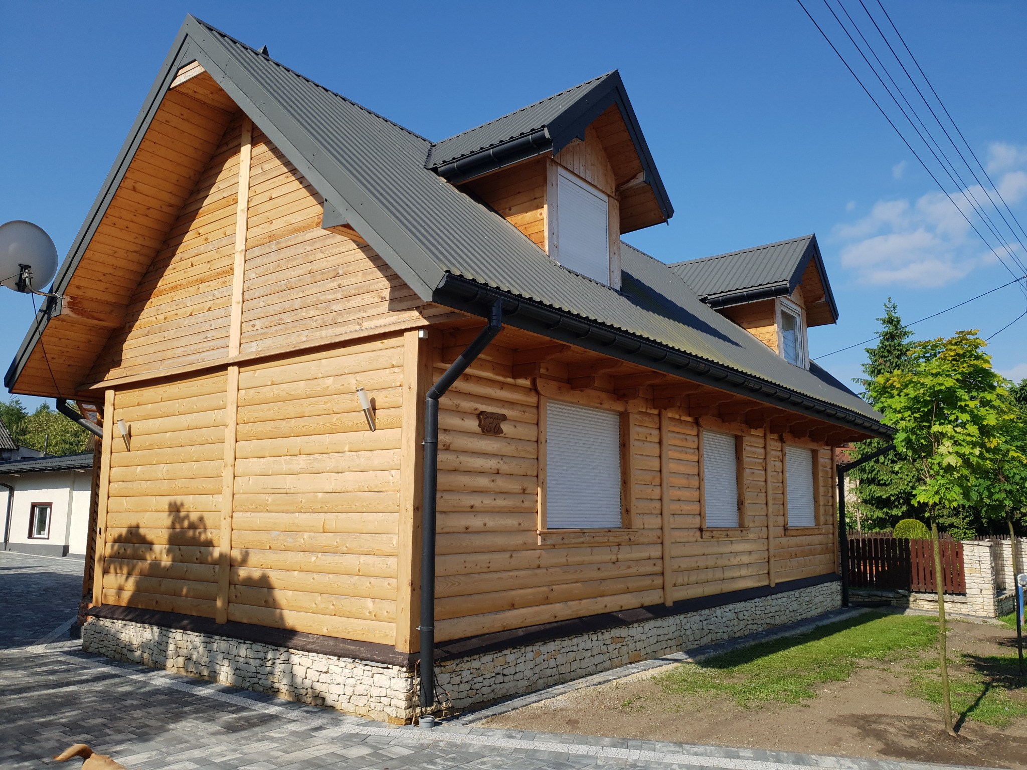 Nowoczesny dom z bali, jasne drewno, ciemny dach z blachy trapezowej, okna z roletami zewnętrznymi, widok z zewnątrz, kostka brukowa, podmurówka z kamienia.