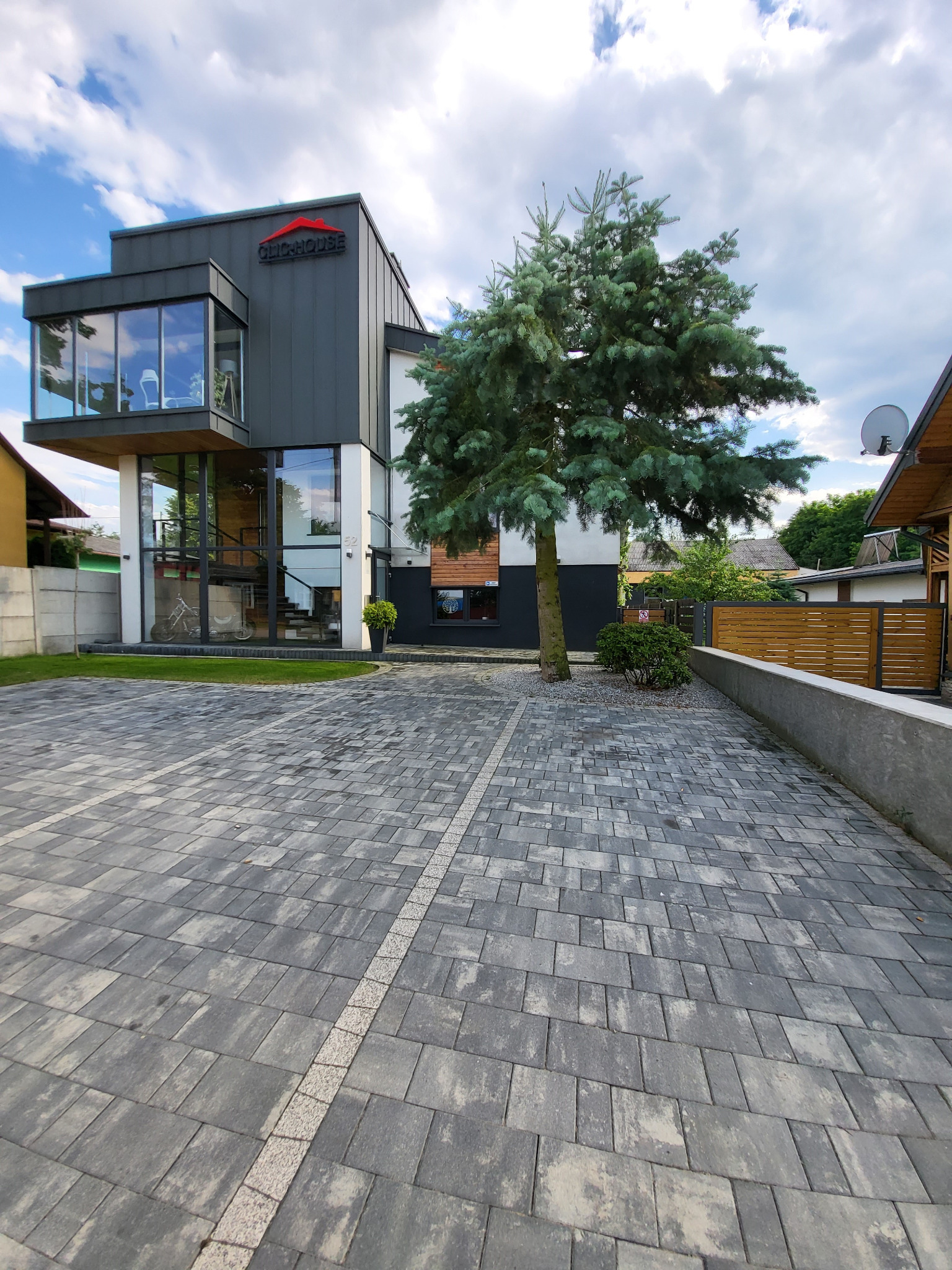 Nowoczesny dom szkieletowy z grafitową elewacją, przeszklonym narożnikiem i logo firmy. Brukowany podjazd i iglaste drzewo na pierwszym planie.