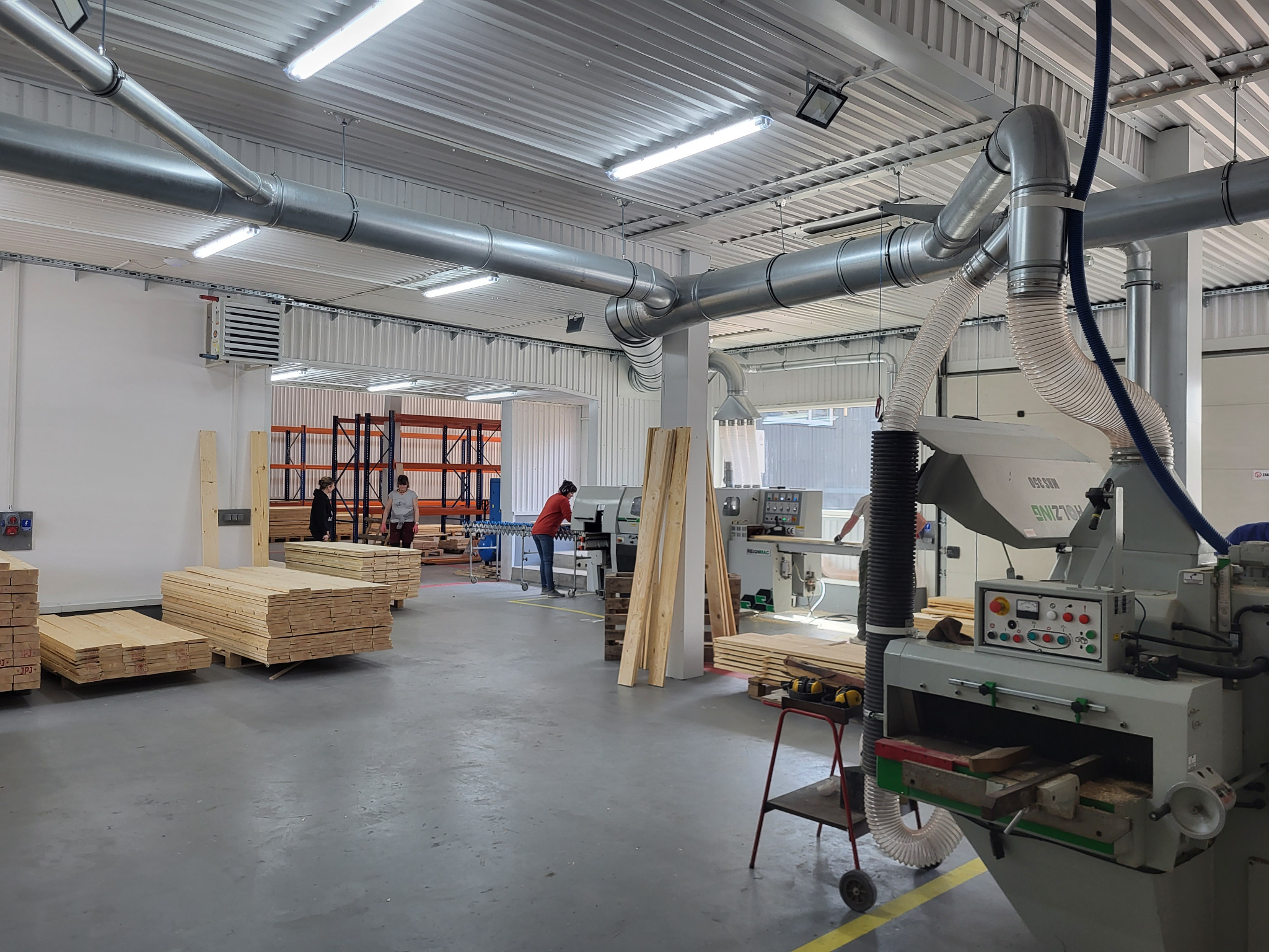 Wnętrze hali produkcyjnej z maszynami do obróbki drewna, ułożonymi deskami i pracownikami w tle. Widoczne rury wentylacyjne pod sufitem.
