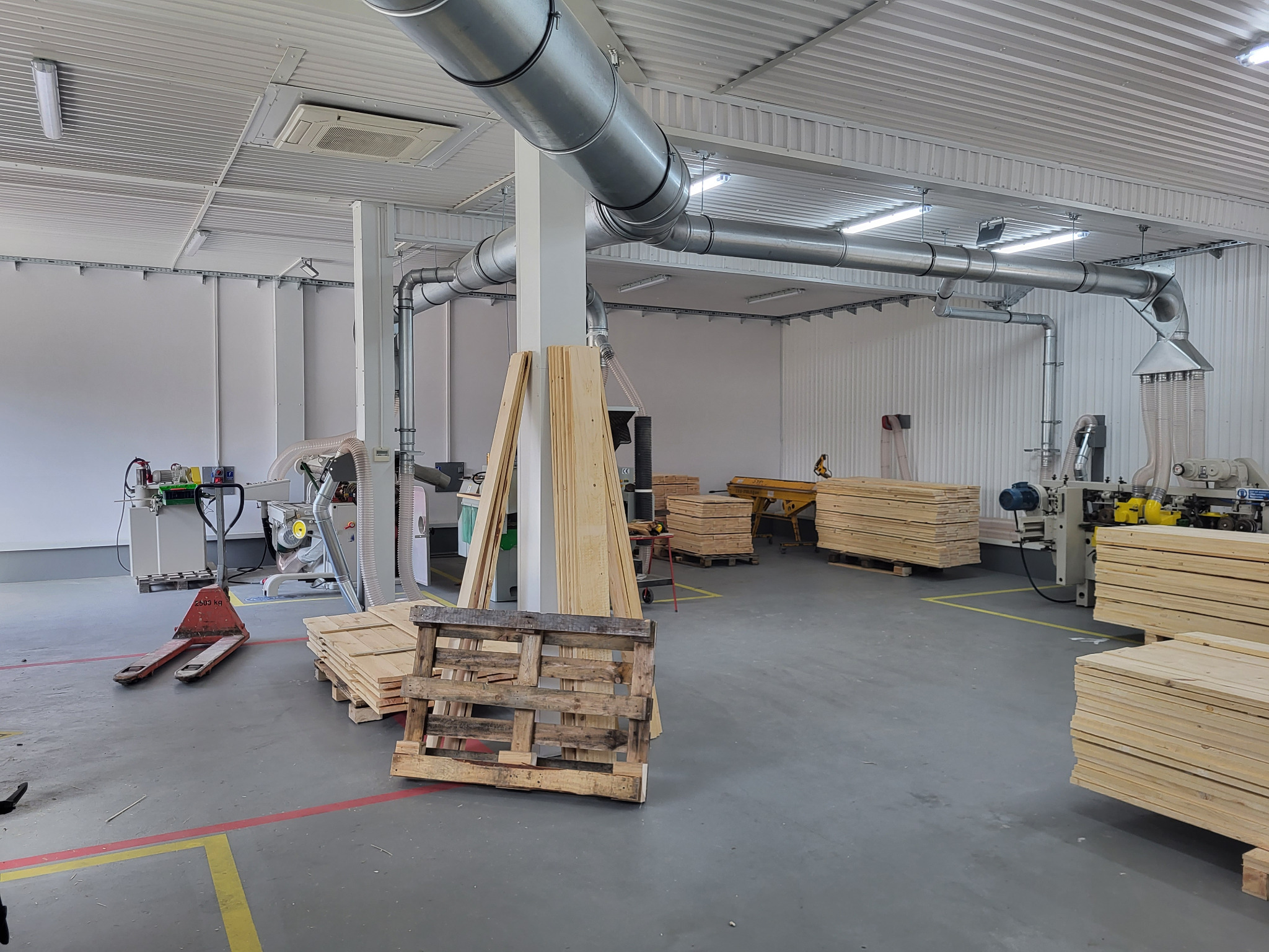 Wnętrze warsztatu stolarskiego z maszynami do obróbki drewna, paletami i ułożonymi deskami. System wentylacji pod sufitem. Widok ogólny, jasne oświetlenie.