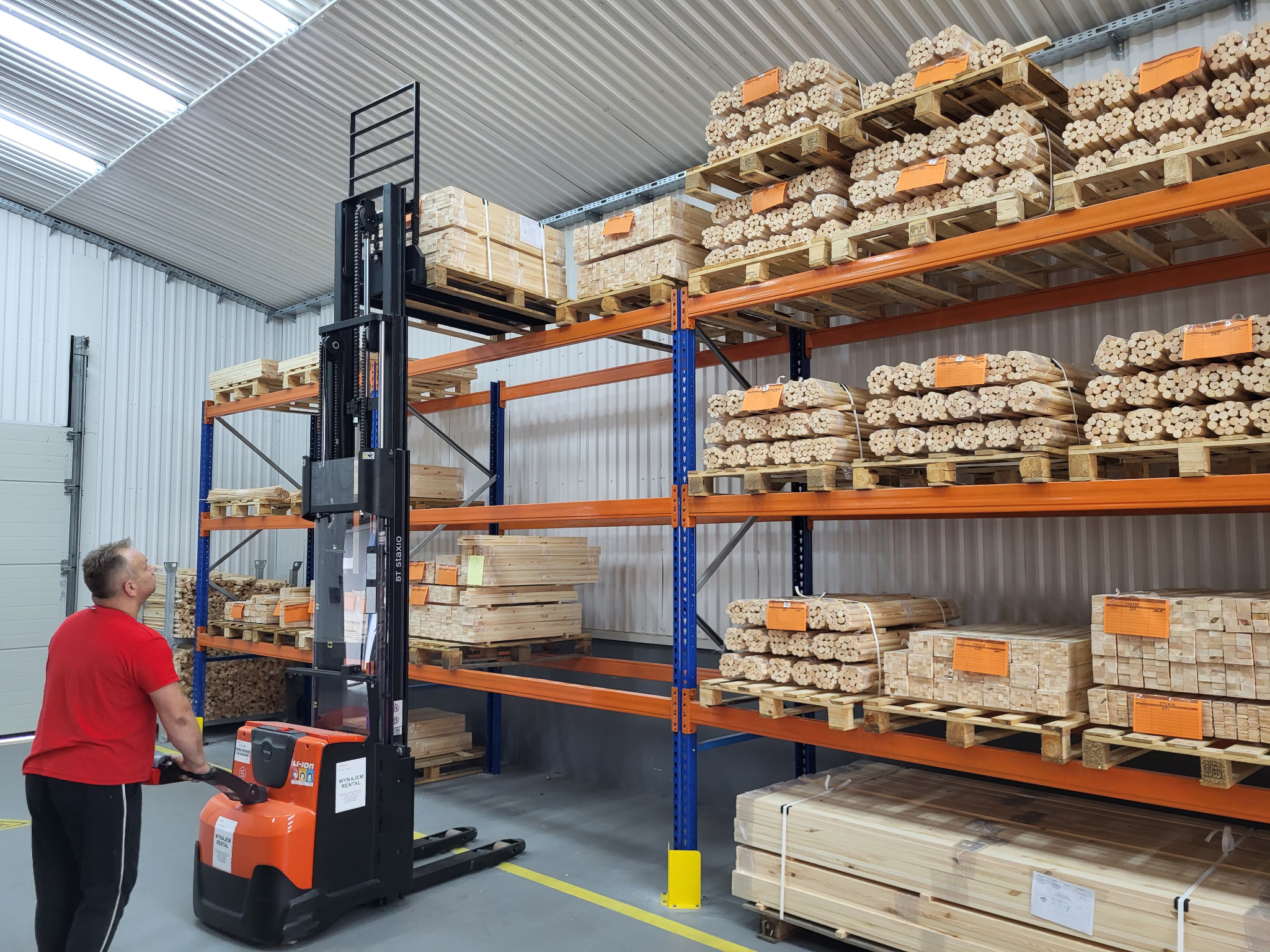 W magazynie operator wózka widłowego podnosi paletę z drewnianymi elementami na regał. Widoczne zapasy materiałów do produkcji drewnianych konstrukcji.