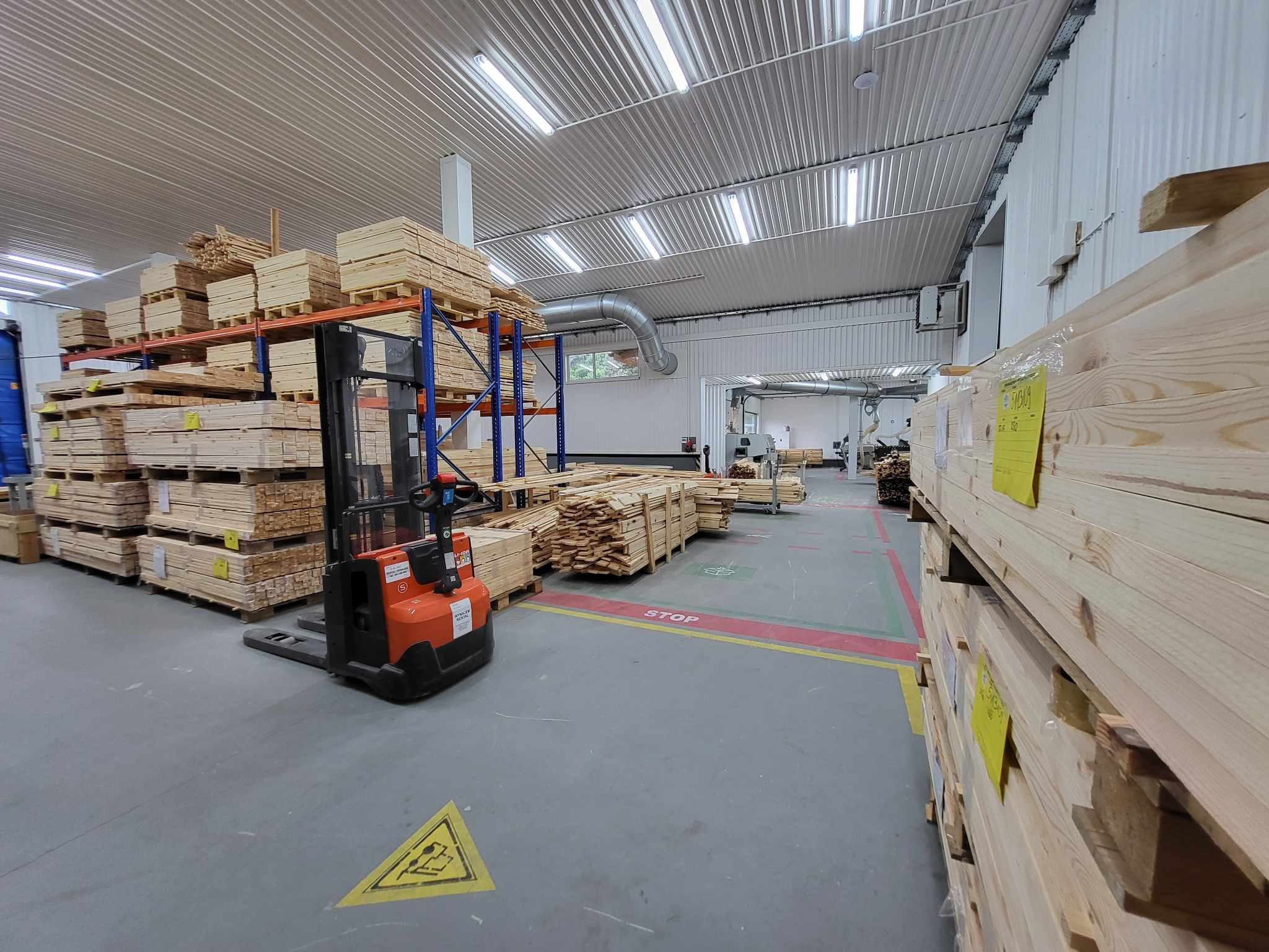 Wnętrze hali produkcyjnej z wózkiem widłowym i ułożonymi paletami z drewnem. Widoczne regały z deskami i elementami drewnianymi. Oświetlenie jarzeniowe.