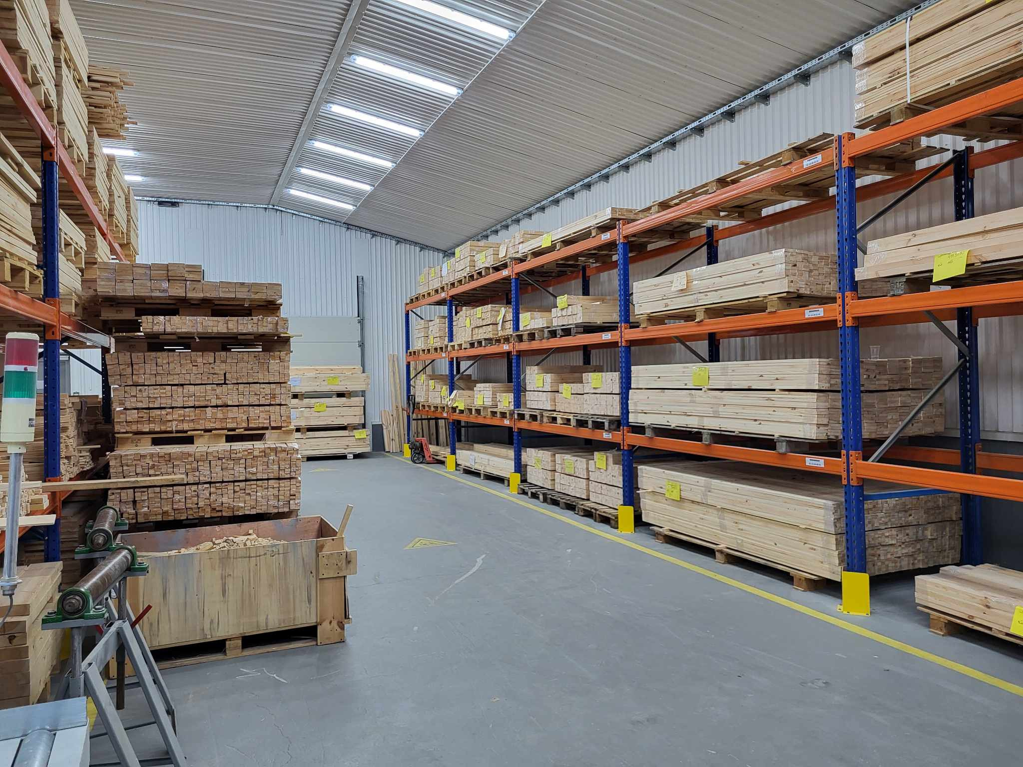 Magazyn tarcicy: rzędy półek z belkami drewna na paletach, w tle maszyna do obróbki drewna i skrzynia z odpadami. Hala z oświetleniem sufitowym.