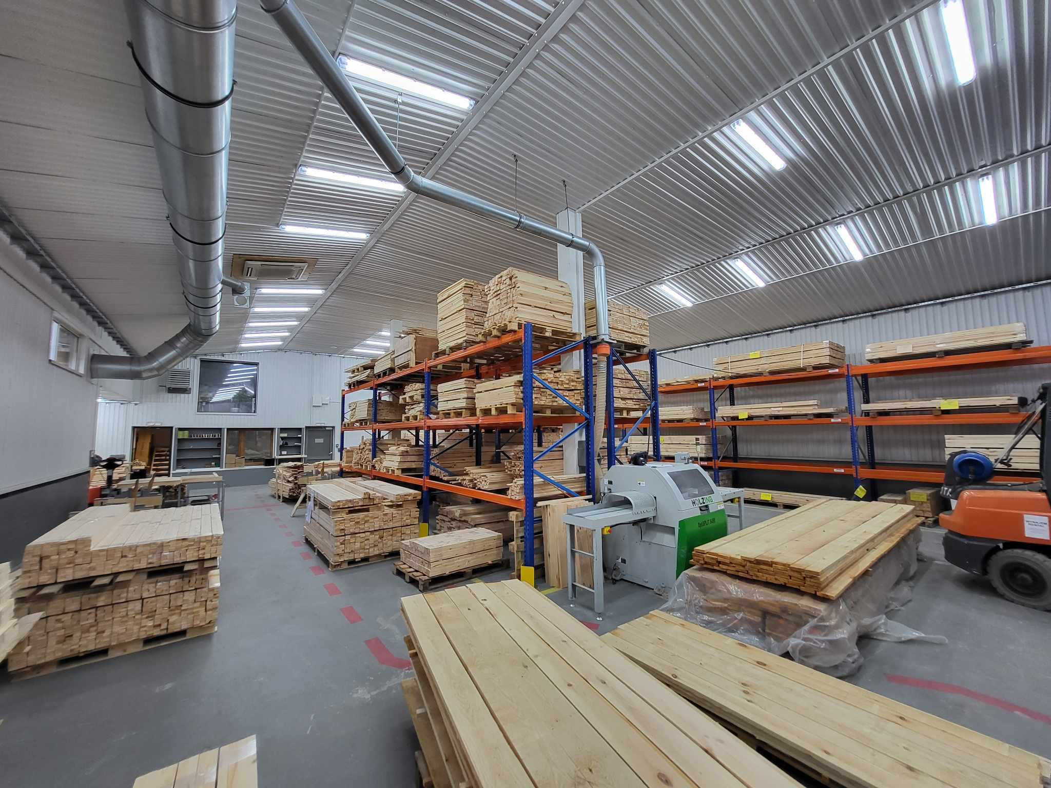 Magazyn drewna z maszynami do obróbki, regały z deskami i wózkiem widłowym. Widoczne elementy wentylacji pod sufitem.