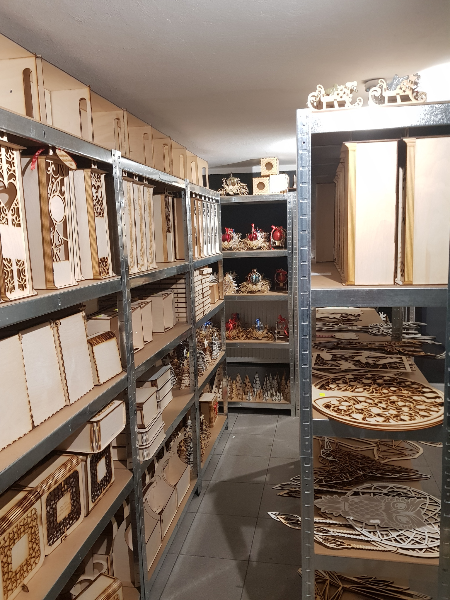 Metalowe regały wypełnione różnorodnymi, surowymi elementami i konstrukcjami z jasnego drewna, niektóre z ażurowymi wycięciami, inne w formie pudełek i ozdób świątecznych.