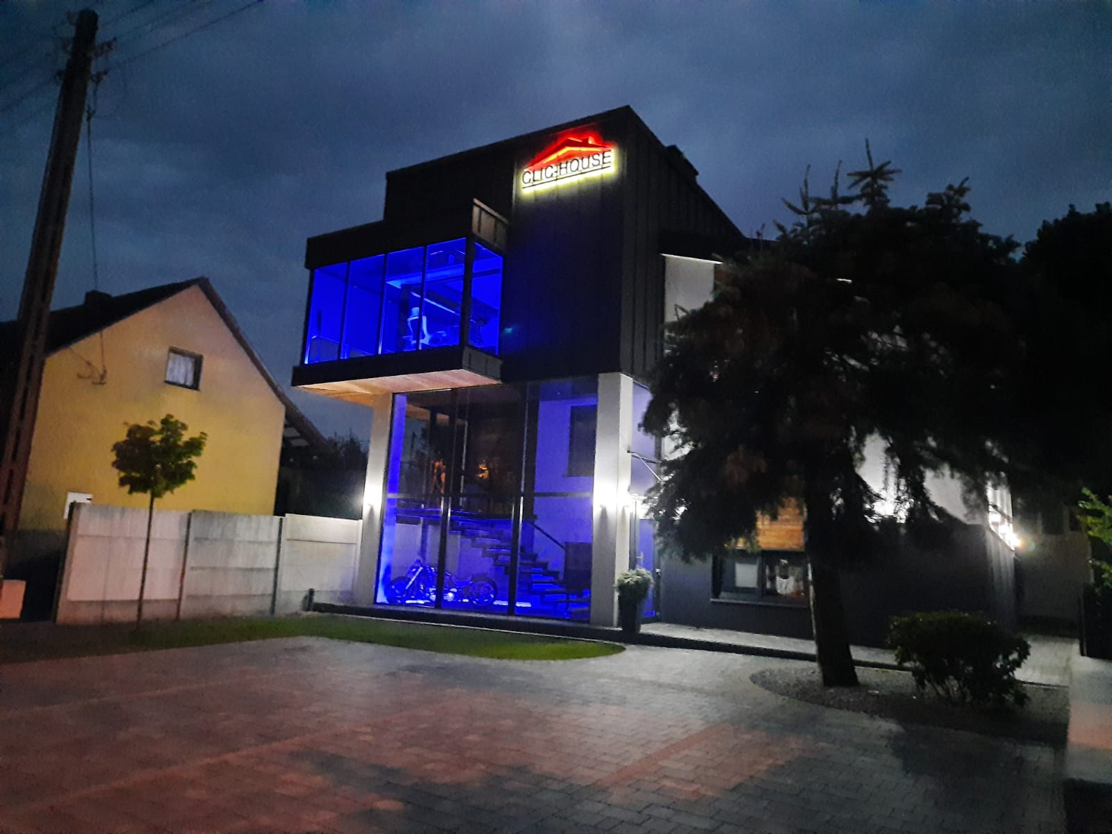 Nowoczesny dom z przeszkloną fasadą i podświetleniem LED w kolorze niebieskim, widoczny z zewnątrz nocą, z logo firmy na dachu.