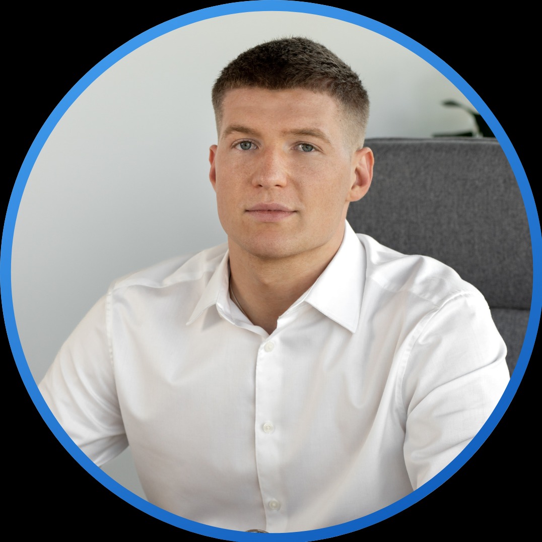 Portret młodego mężczyzny w białej koszuli, siedzącego na szarym krześle, na neutralnym tle, w niebieskim okręgu kadrującym.