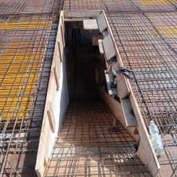 Dan Bud - Zbrojenie stropu z otworem na schody, widoczne deski szalunkowe, siatka zbrojeniowa, narzędzia pracy i butelka wody. Prace budowlane w toku, widok z góry.