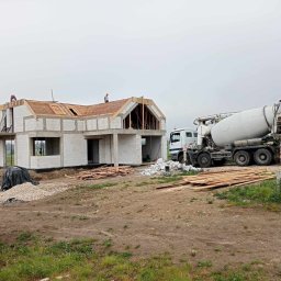 Dan Bud - Budowa domu jednorodzinnego z betonu komórkowego. Na dachu dwóch robotników, obok betoniarka. Widoczne materiały budowlane i teren budowy.