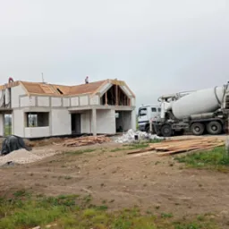 Budowa domu jednorodzinnego z betonu komórkowego. Na dachu dwóch robotników, obok betoniarka. Widoczne materiały budowlane i teren budowy.