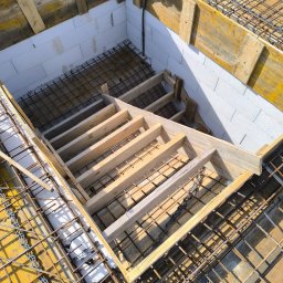 Dan Bud - Szkielet betonowych schodów z drewnianym szalunkiem i zbrojeniem, widok z góry. W tle białe bloczki, a na obrzeżach żółte deski szalunkowe i narzędzie.