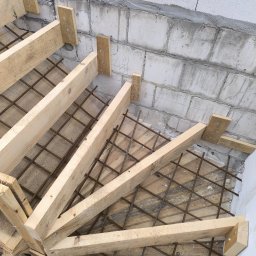 Dan Bud - Szczegółowy widok z góry na szalunek i zbrojenie pod betonowe schody, przymocowane do ściany z bloczków betonowych. Widoczne elementy drewniane i metalowe.