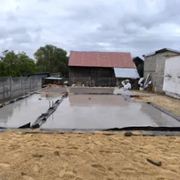 Wylany fundament pod budynek w Sobkowie. Widoczna czarna folia, mokry beton i piasek. W tle budynki gospodarcze i ogrodzenie.