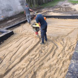 Dan Bud - Prace ziemne: ubijanie piasku zagęszczarką w przygotowanym wykopie pod fundament. Widoczny mur oporowy i drugi pracownik w tle.